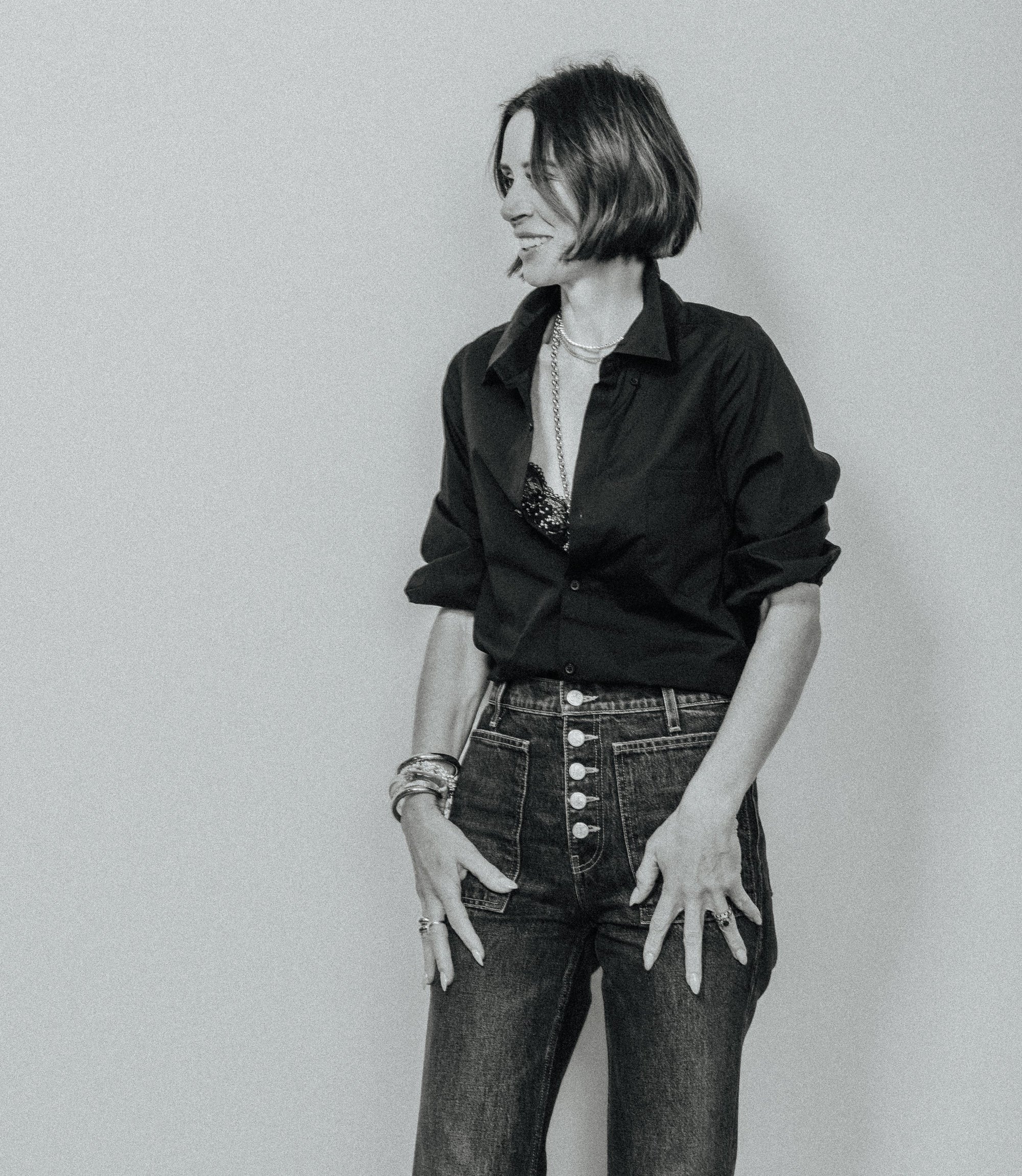 A woman with short dark hair stands against a plain light gray wall, wearing a black button-front shirt, dark high-waisted jeans with prominent front buttons, layered necklaces, and bracelets.
